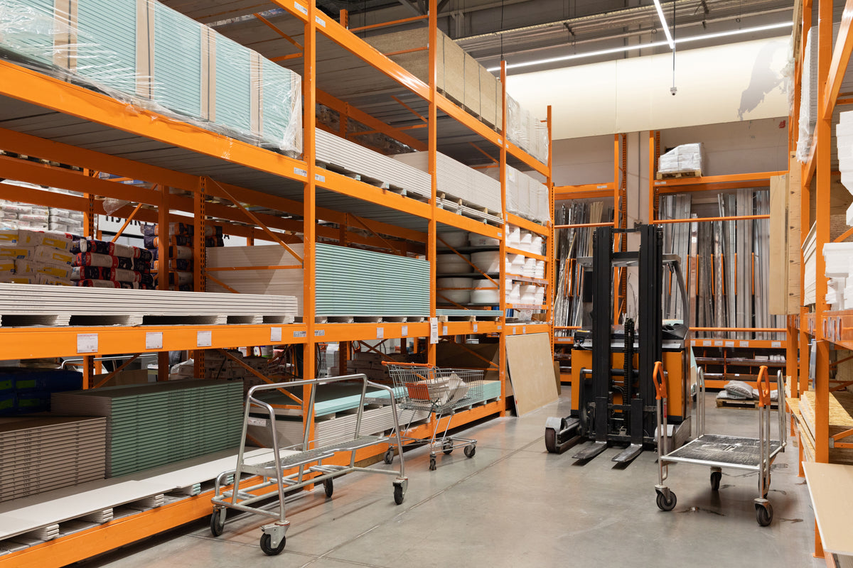 Lagergang mit orangefarbenen Regalen, Transportwagen und Stapeln von Baumaterialien.